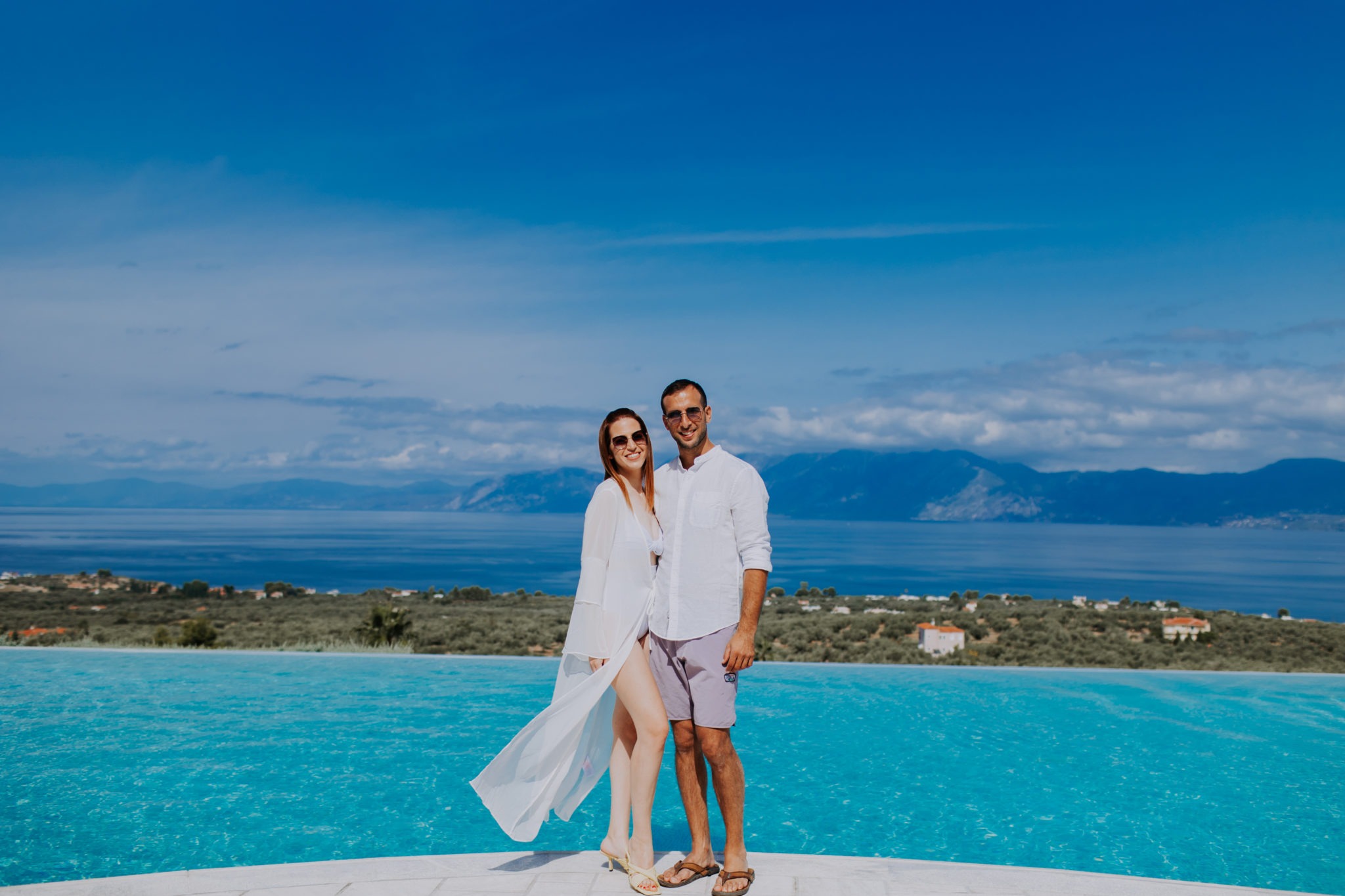Couple Shani & Sean in white wedding attire by an infinity pool overlooking the blue sea in Greece, featured in Sirtaki Events' couple stories.