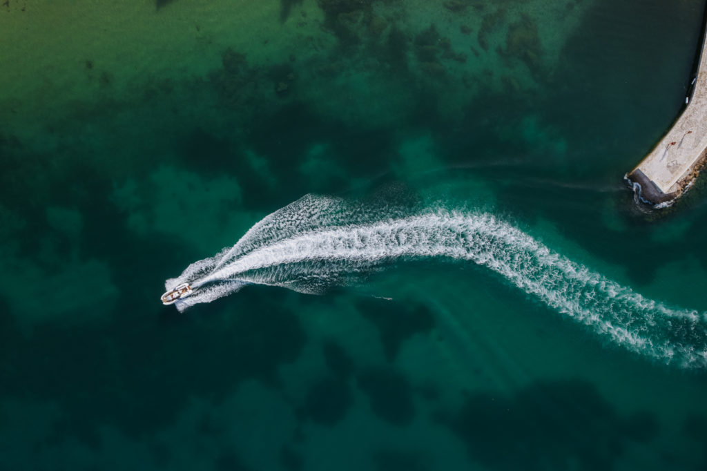 An aerial drone shot of a luxury speed boat cruising through crystal-clear turquoise waters, leaving a long white foam wake in the sea during a private celebration in Greece.